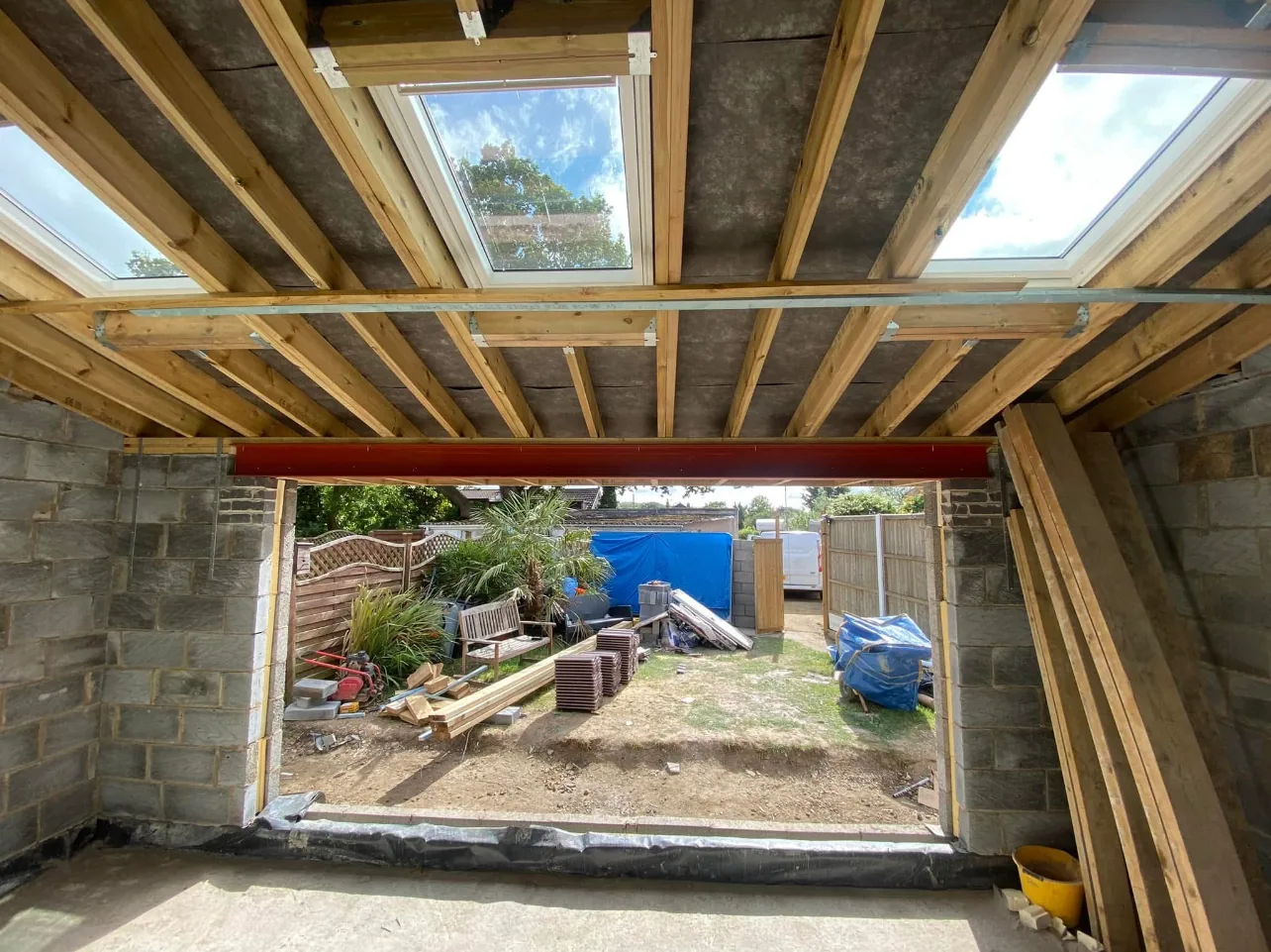 Inside a new extension shell: timber roof joists, three rooflights and a red steel beam spanning the wide rear opening