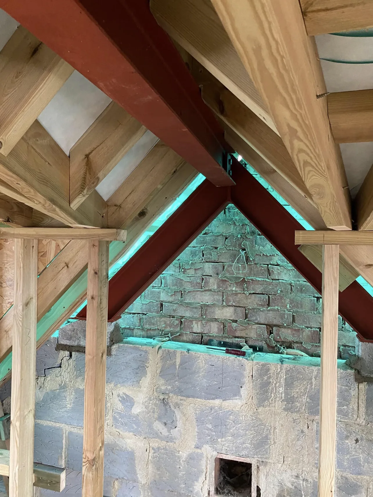 Inside a roof under alteration: red structural steels meeting at the ridge, new timber rafters, and temporary props below